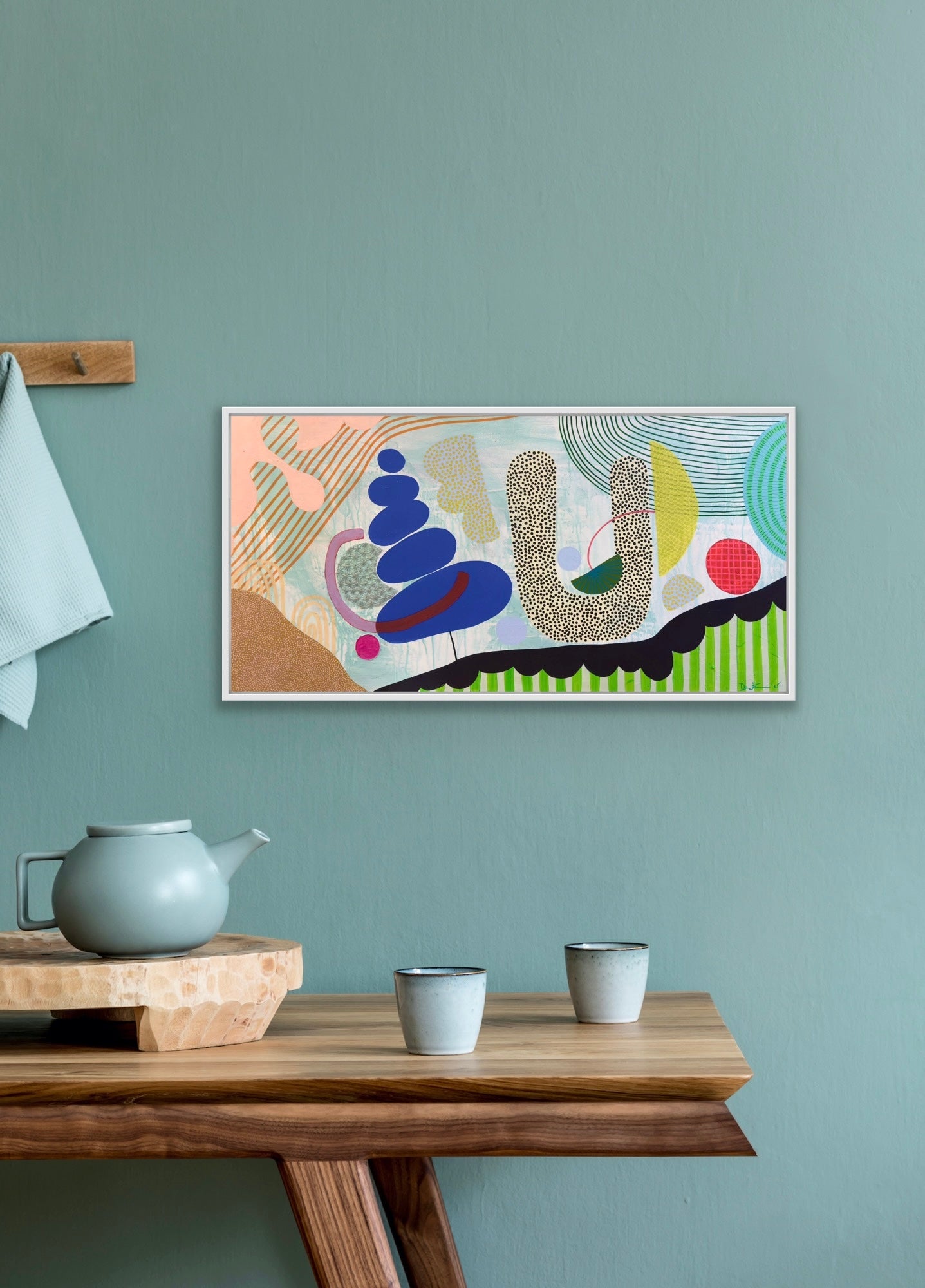Abstract artwork on a wall above a wooden table with a teapot and cups.